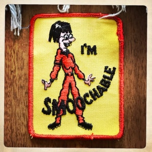 May include: A yellow embroidered patch with a cartoon figure wearing a red jumpsuit and glasses. The figure is standing with the word "I'm Smoachable" written around it.