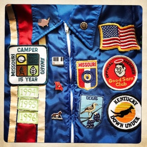 Könnte beinhalten: Blaue Satinjacke mit weißem Reißverschluss und verschiedenen Aufnähern. Zu den Aufnähern gehören die amerikanische Flagge, "Missouri Camper Award 15 Year", "Good Sam Club", "Texas" und "Kentucky Down Under". Die Jacke hat auch Anstecknadeln und die Jahre 1994, 1995 und 1996.