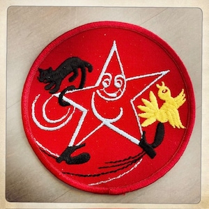 May include: A red circular patch with a white star in the center. The star has a smiling face and is holding a yellow bird with black legs and a black cat with white eyes. The background is red with white stitching.