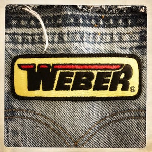Può includere: Una toppa ricamata gialla e nera con la parola "WEBER" in lettere nere e una linea rossa sopra la parola. La toppa è cucita su un tessuto di jeans blu.