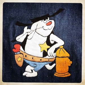 May include: Embroidered patch featuring a cartoon character in a cowboy hat and vest, leaning on a yellow fire hydrant. The character is white with black and red accents, wearing a sheriff's star and blue shorts.
