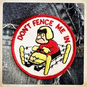 May include: A white embroidered patch with a red border. The patch features a cartoon character wearing a red jacket and a yellow helmet, riding a yellow snowmobile through a barbed wire fence. The text "DON'T FENCE ME IN" is written around the character.