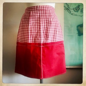 May include: A red and white gingham apron with a solid red bottom. The apron has a white seam around the bottom edge.