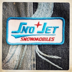 Puede incluir: Un parche bordado azul y blanco con el texto "Sno-Jet Snowmobiles" en un estilo retro.