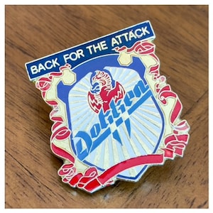 May include: A blue and red enamel pin with a shield design. The shield features a red phoenix, a white background with gold rays, and the word "Dokken" in blue. The shield is surrounded by red and gold ribbons. The text "Back for the Attack" is printed in white on a blue banner above the shield.