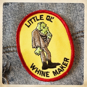 May include: A yellow and red embroidered patch with a cartoon monster in a suit. The text on the patch reads "Little Qu' Whine Maker!"