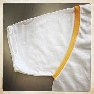Puede incluir: Camiseta blanca con ribete amarillo alrededor del cuello y las mangas.