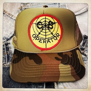 Puede incluir: Una gorra de camionero de camuflaje verde con un parche bordado amarillo y rojo que dice "Operator" con un diseño de diana.