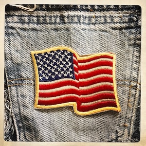 Può includere: Un piccolo patch della bandiera americana cucito su un pezzo di tessuto denim blu. La bandiera è rossa, bianca e blu con un bordo giallo.