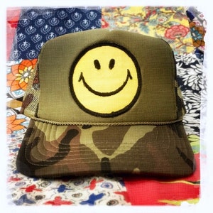 May include: A green camouflage trucker hat with a yellow smiley face patch. The hat has a mesh back and a black brim.