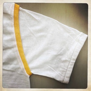 Puede incluir: Camiseta blanca con ribete amarillo alrededor del cuello y las mangas.