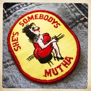 Pode incluir: Um remendo bordado amarelo e vermelho com uma mulher de desenho animado com um vestido vermelho sentada em uma cadeira. O texto ao redor da mulher diz "She's Somebody's Mutha".