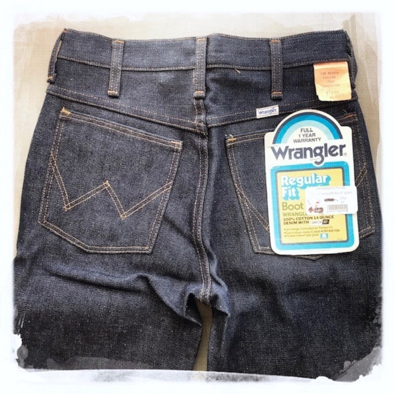 May include: A pair of dark blue denim jeans with a back pocket featuring a stitched 'W' logo. The jeans have a label on the back pocket that reads 'Wrangler Regular Fit Boot Wrangler 100% Cotton 14 Ounce Denim with'.