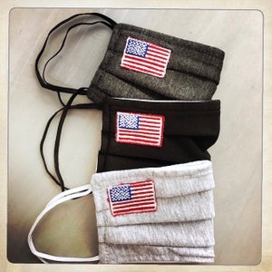 May include: Three face masks in shades of gray, black, and light gray, each adorned with a small embroidered American flag patch. The masks have pleated designs and black or white elastic ear loops. The masks are stacked on top of each other.