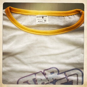 Puede incluir: Camiseta blanca con un ribete amarillo alrededor del cuello. La camiseta tiene una impresi&oacute;n descolorida de "STAR" en la parte delantera. La etiqueta de la camiseta dice "Medallist Pride" y "Adult M".
