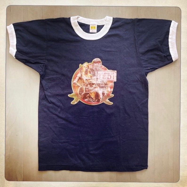 Op de afbeelding: Een donkerblauw t-shirt met een witte boord. Het shirt heeft een vintage afbeelding van een sc&egrave;ne uit de film "Return of the Jedi" met de tekst "Return of the Jedi" op een bord.