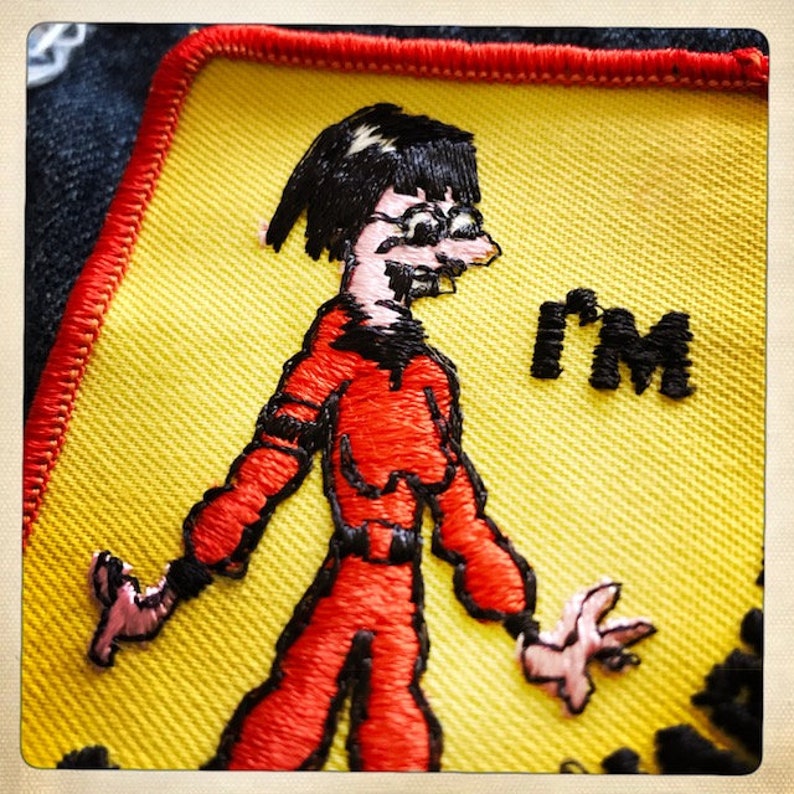 May include: A yellow embroidered patch with a cartoon figure wearing a red jumpsuit and black boots. The figure has black hair and glasses. The text "I'M" is embroidered in black on the patch.