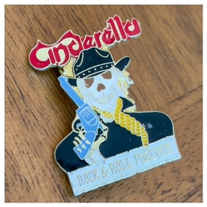 Könnte beinhalten: Eine goldene und rote Emaille-Anstecknadel mit einem Skelett, das einen Cowboyhut trägt und eine Pistole hält. Die Anstecknadel trägt die Aufschrift "Cinderella" und "Rock & Roll Forever".