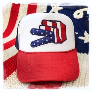 May include: A white and red trucker hat with a peace sign embroidered on the front. The peace sign is in the colors of the American flag. The hat has a red mesh back.