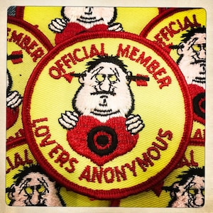 May include: Yellow and red embroidered patch featuring a cartoon character holding a heart with a target in the centre. The text reads "Official Member Lovers Anonymous".