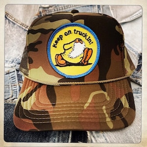 May include: Camouflage trucker hat with a yellow and blue patch that says "keep on truckin!" and features a cartoon character.