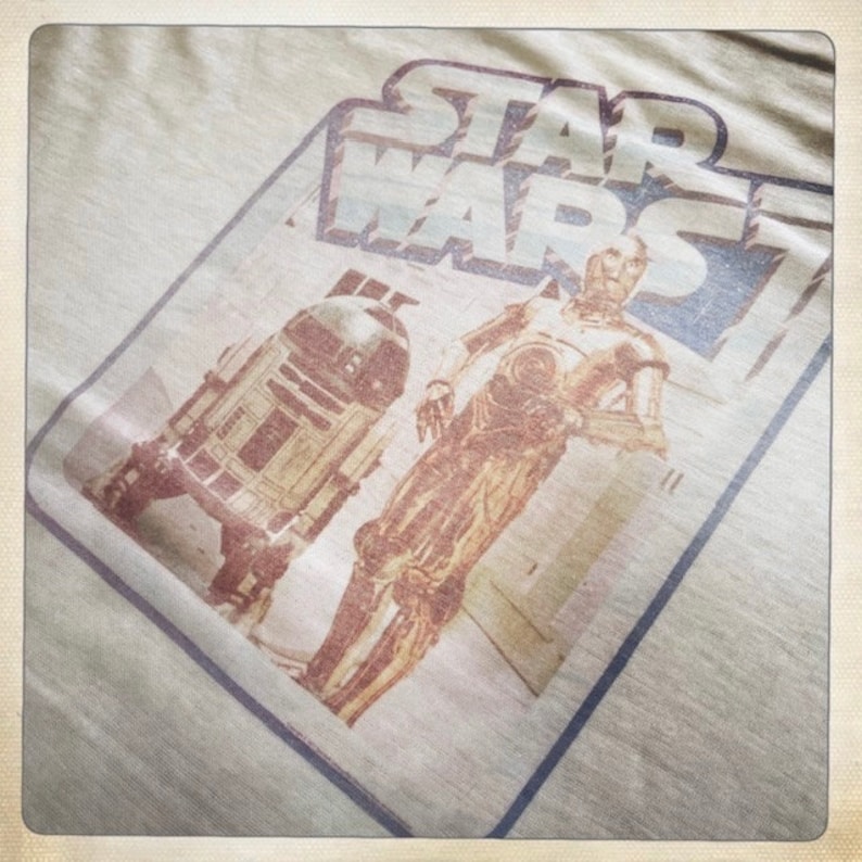 Puede incluir: Una camiseta vintage de Star Wars con una imagen descolorida de R2-D2 y C-3PO. La camiseta es beige y tiene un borde azul y blanco alrededor de la imagen.