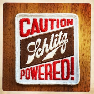 May include: White embroidered patch with red text that reads "CAUTION Schlitz POWERED!" The word "Schlitz" is in a brown cursive font.