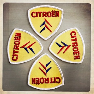 Może przedstawiać: Cztery żółte, trójkątne łatki z tkaniny z białymi szwami wokół krawędzi. Każda łatka ma napis "CITROËN" czerwonymi literami i stylizowany wzór w chevron w kolorze czerwonym i niebieskim.