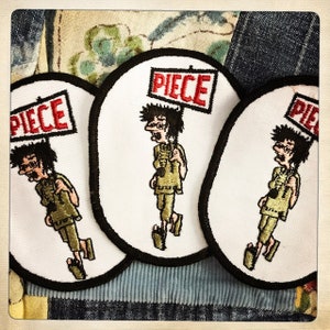 May include: Three white embroidered patches with black outlines, each featuring a cartoon character holding a sign that says "PIECE". The character is wearing a green shirt and brown trousers.