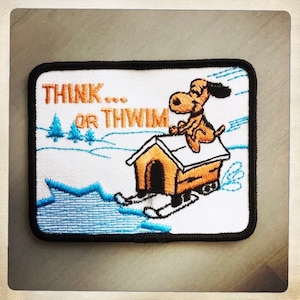 May include: A white embroidered patch with a cartoon dog sitting on top of a dog house on skis. The text "THINK... OR THWIM" is above the dog house.