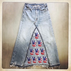 Op de afbeelding: Een lange denim rok met een driehoekig patroon van vredestekens. Elk vredesteken is versierd met de Amerikaanse vlag.