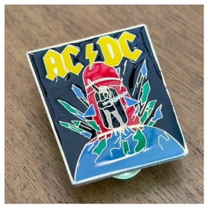 May include: A black and yellow enamel pin with the text "AC/DC" in a lightning bolt design. The pin features a red rocket ship with a figure in black standing on top of the rocket, which is launching from a blue and green globe.