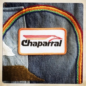 Puede incluir: Una chaqueta vaquera con un dise&ntilde;o bordado de arco&iacute;ris y un parche blanco con el texto "Chaparral" y un logotipo de p&aacute;jaro rojo, naranja y amarillo.