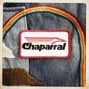 Puede incluir: Una chaqueta vaquera con un parche bordado de arco&iacute;ris y un parche bordado de rojo, blanco y negro que dice "Chaparral".