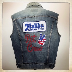May include: A blue denim vest with a red embroidered "Pit Crew" design, a "Malibu Grand Prix" patch, and a peace sign patch with an American flag design.