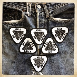 May include: Five white triangular patches with black text that reads "Kiss My Patch" on a pair of blue denim jeans.