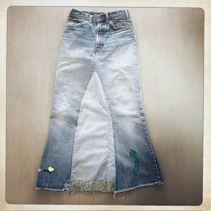 May include: A long denim skirt made from different shades of blue denim. The skirt has a high waist and a flared silhouette. The skirt is decorated with embroidered flowers and a green leaf design.