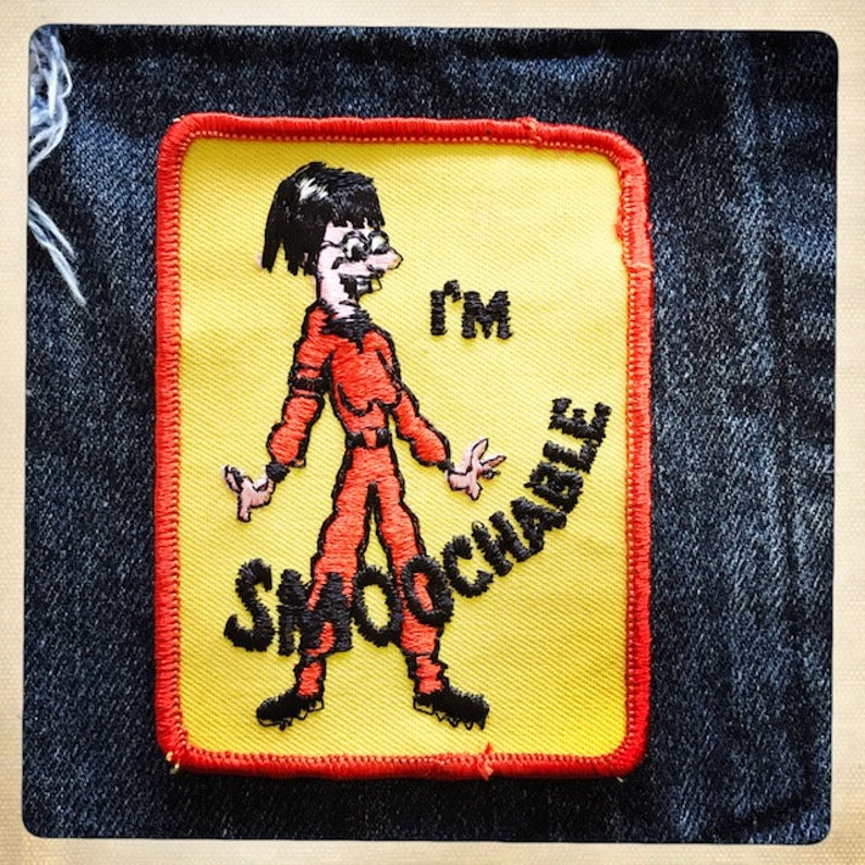 May include: A yellow iron-on patch with a red border. It features a cartoon character wearing a red jumpsuit and glasses. The text "I'm smoochable" is written around the character.