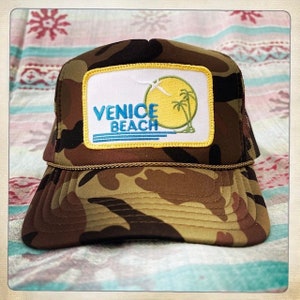 Puede incluir: Una gorra de camionero de camuflaje con un parche amarillo, azul y blanco que dice "Venice Beach". El parche presenta una palmera y un pájaro.
