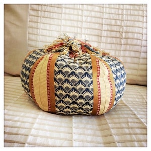 May include: A round, blue and cream patterned cushion with gold and red ribbon accents. The cushion is shaped like a pumpkin.