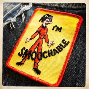May include: A yellow and red embroidered patch with a cartoon figure in a red jumpsuit. The figure is standing with arms outstretched and the text "I'm smoochable" is written in black.