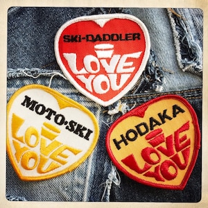 May include: Three heart-shaped patches with the words "Ski-Daddler Love You", "Moto-Ski Love You", and "Hodaka Love You" in yellow and red lettering on a denim background.