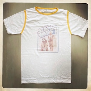 Puede incluir: Camiseta blanca con ribete amarillo alrededor del cuello y las mangas. La camiseta presenta un gr&aacute;fico vintage de Star Wars con R2-D2 y C-3PO.