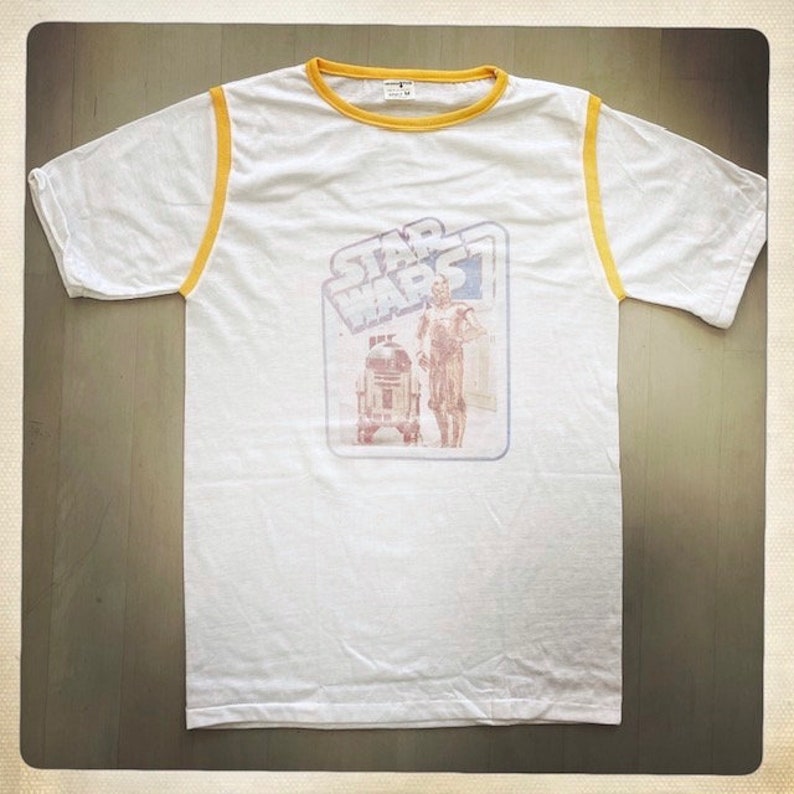 Puede incluir: Camiseta blanca con ribete amarillo alrededor del cuello y las mangas. La camiseta presenta un gr&aacute;fico vintage de Star Wars con R2-D2 y C-3PO.