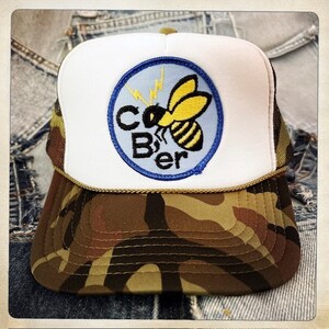 May include: A white and camo trucker hat with a blue and white patch featuring a yellow bee with black stripes and the text "C-B'er".