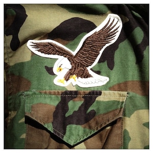 May include: A camouflage fabric jacket with a white and brown embroidered eagle patch on the left chest pocket.