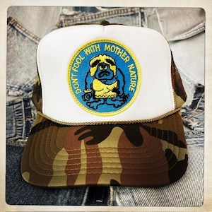 May include: A white and camo trucker hat with a yellow, blue, and white embroidered patch that says "Don't Fool With Mother Nature". The patch features a cartoon character with a yellow shirt and blue pants.