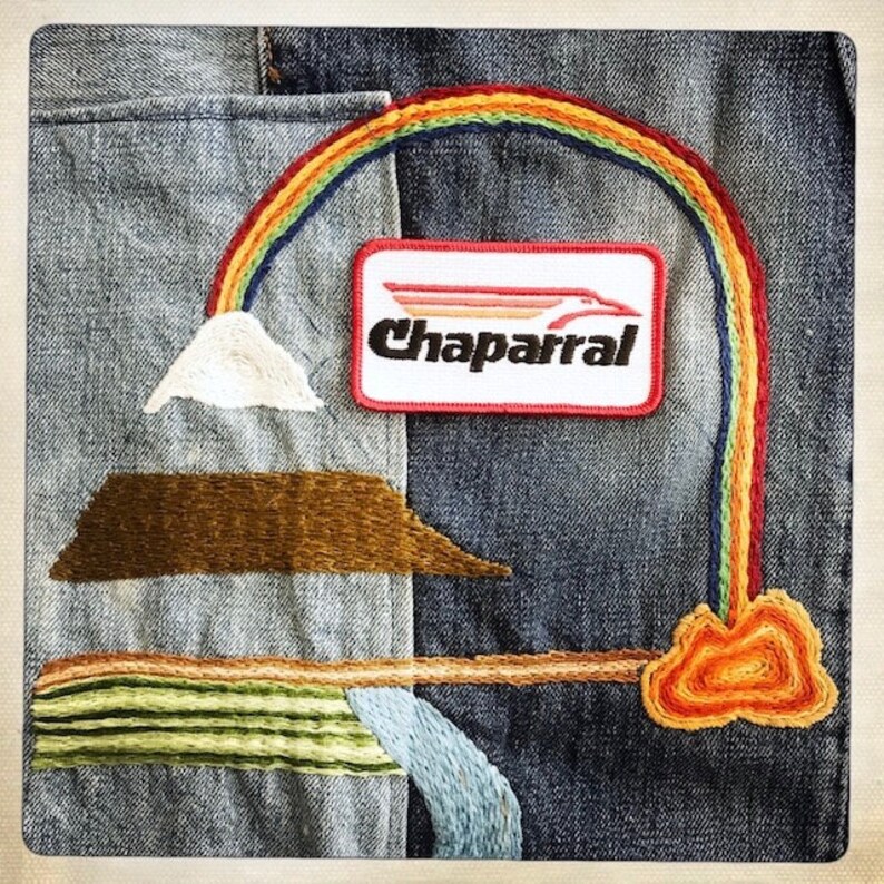 Puede incluir: Una chaqueta vaquera con detalles bordados, incluyendo un arco&iacute;ris, una monta&ntilde;a, una nube y un parche que dice "Chaparral".