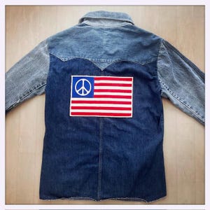Könnte beinhalten: Eine langärmelige Jeansjacke mit zweifarbiger Waschung, hellblauem Sattel und Ärmeln sowie einem dunkelblauen Rumpf. Auf dem Rücken befindet sich ein rechteckiger Aufnäher mit einer rot-weiß gestreiften Flagge und einem blauen Friedenssymbol.