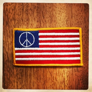 May include: A small embroidered patch with a peace sign in the top left corner and an American flag design in red, white, and blue.